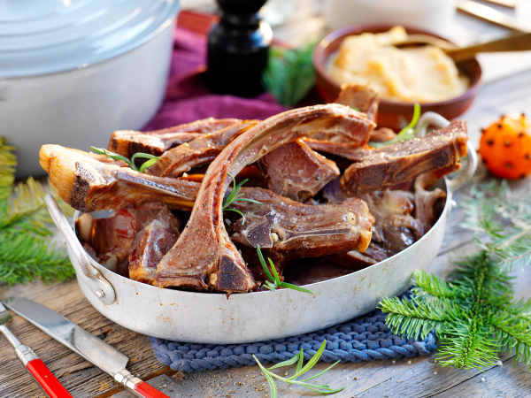 Pinnekjøtt i en hvit serveringsskål med grønne urter og tilbehør rundt på et bord.