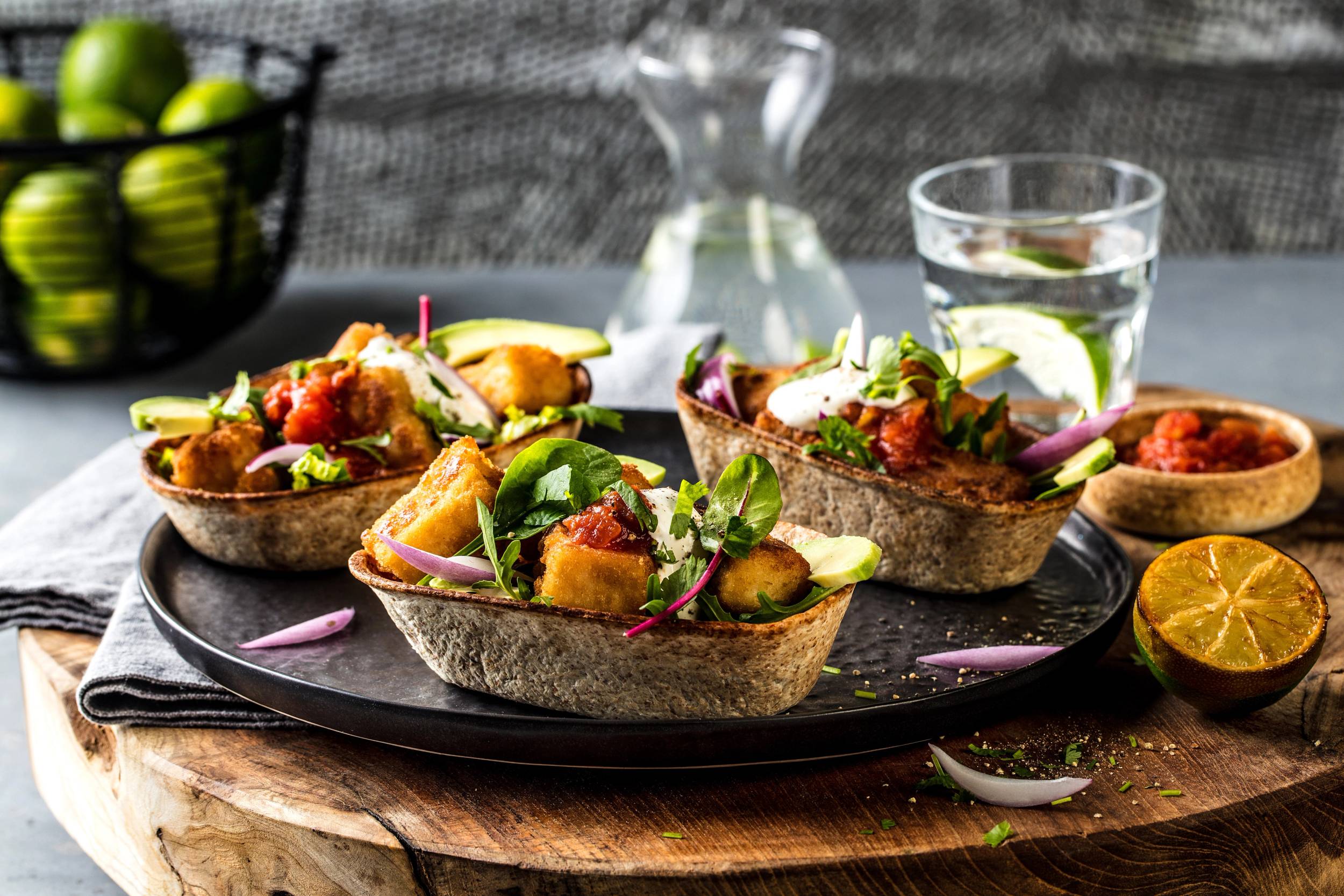 Fisketacos med fisk, grønnsaker og lime på en svart tallerken, med en glass vann og en karaffel i bakgrunnen.