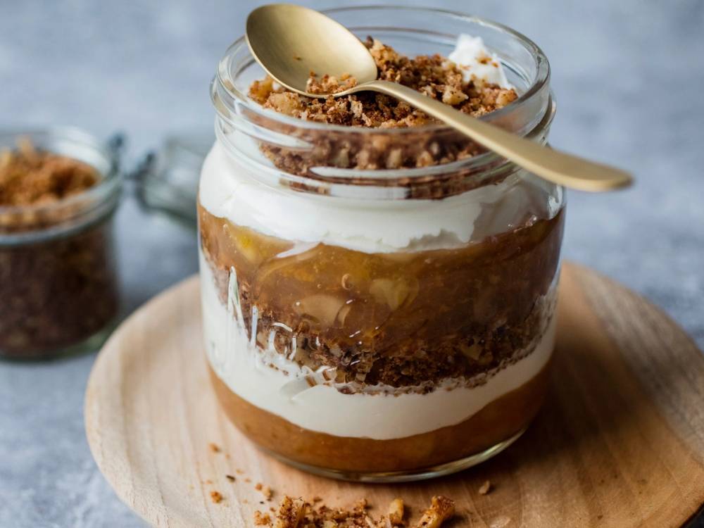 En lagdelt dessert i et glass med krem, sjokoladekrønsj og en skje på toppen, plassert på et rundt trebrett.