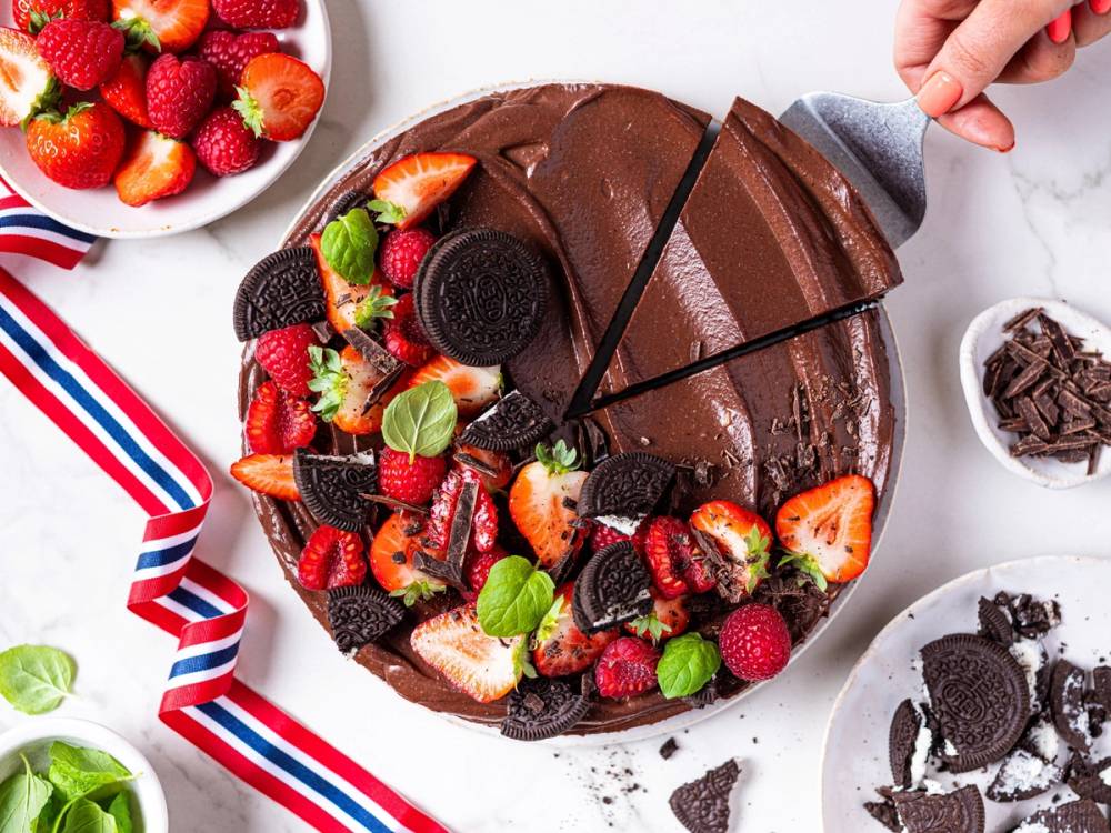 En sjokolademousse-kake pyntet med friske jordbær, bringebær, og Oreo-kjeks, med en person som skjærer i den.