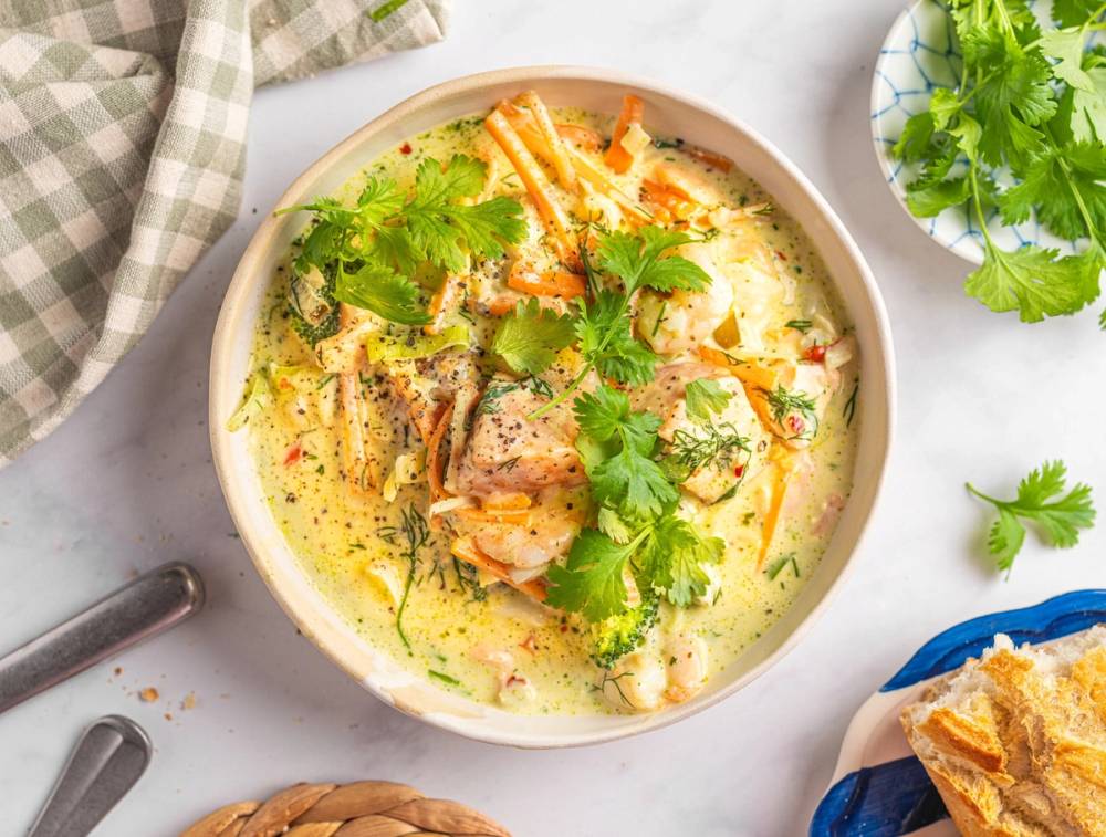 En skål med fiskesuppe med grønnsaker og urter, servert på et hvitt bord med en grønn plante i bakgrunnen.