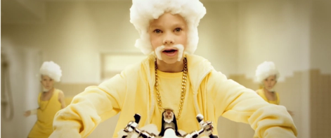 En person vasker hendene på et offentlig toalett, iført gul jakke og smykker, med flere andre personer i bakgrunnen.