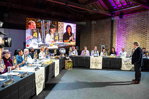 En gruppe mennesker deltar i en konkurranse eller quiz, med store skjermer som viser deltakerne og en dommer foran en lang bord med utstyr og mat, i et rom med brune murvegger og teppebelagt gulv.