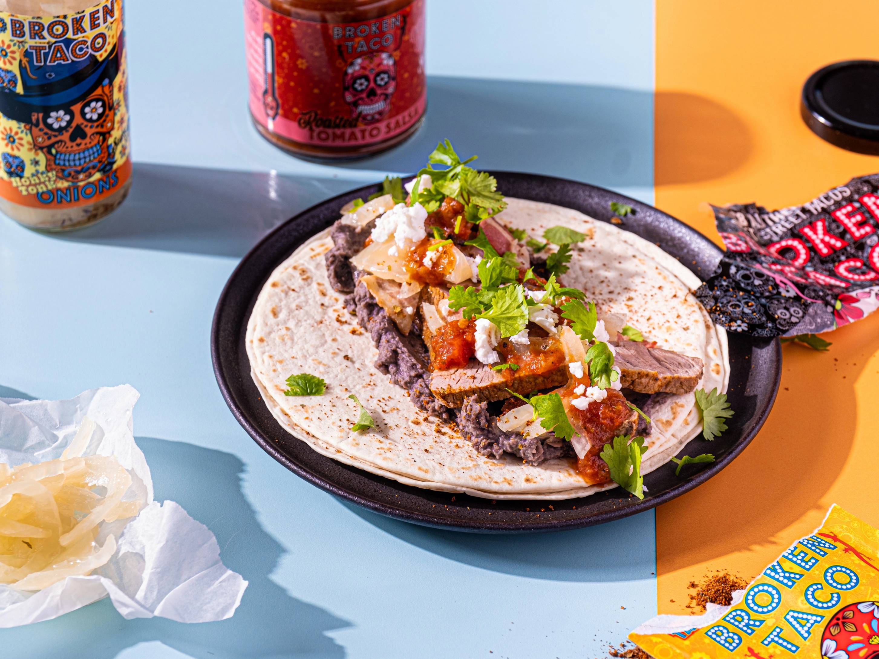 Tacos med kjøtt, salsa, løk og koriander på en svart tallerken, omgitt av brus og snacks på et fargerikt bord.