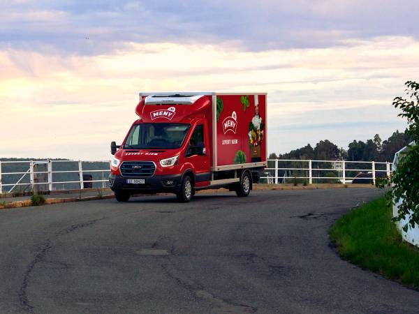 En rød matleveringsbil med Meny-logo kjører på en vei langs sjøen eller innsjøen med en hvit hekk og trær i bakgrunnen.