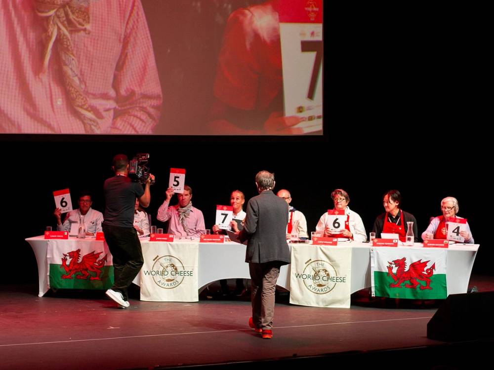 Dommere vurderer bedømming under VM 2022, med en stor skjerm i bakgrunnen og deltakere som sitter ved bord med Wales-flagger.