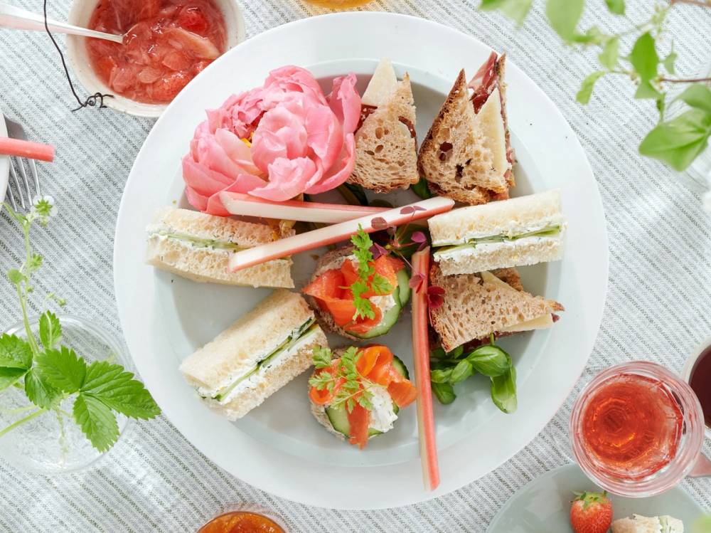 Et serveringsfat med små sandwicher, kaker og frukt, plassert på et bord dekket med en hvit duk og omgitt av grønne blader og glass med rosa drikke.