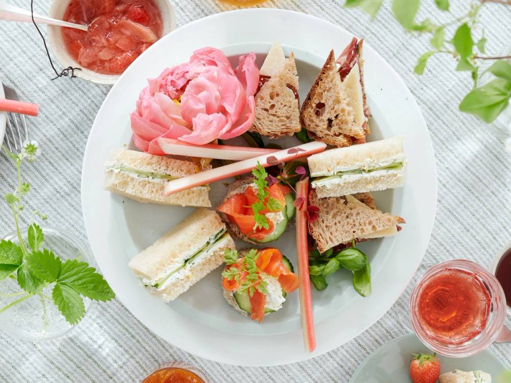 Et serveringsfat med små sandwicher, kaker og frukt, plassert på et bord dekket med en hvit duk og omgitt av grønne blader og glass med rosa drikke.