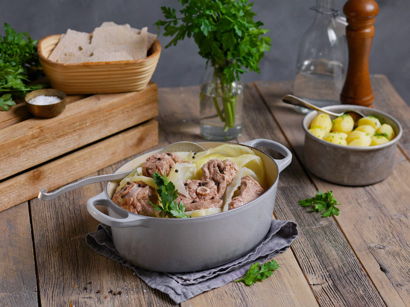 Fårikål i en stor gryte med kjøtt og kål, omgitt av poteter og urter på et kjøkkenbord.