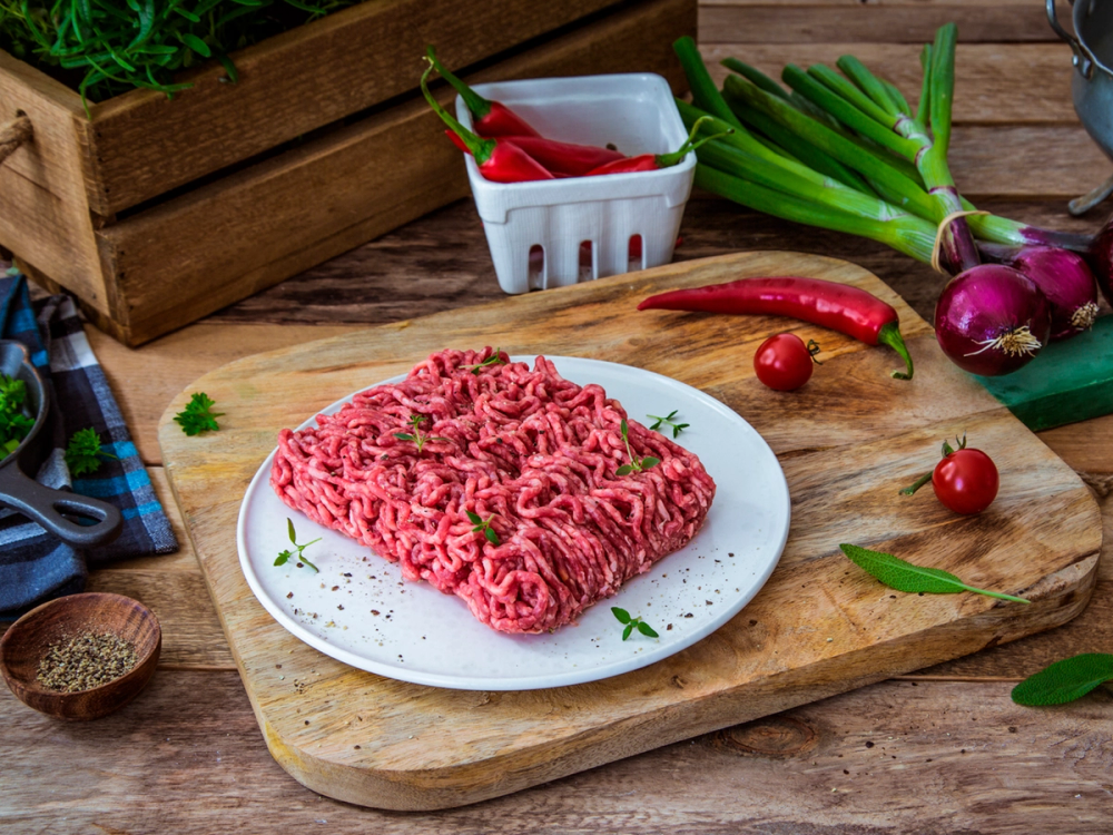 Kjøttdeig på en hvit tallerken med grønnsaker og krydder rundt på et kjøkkenbord.