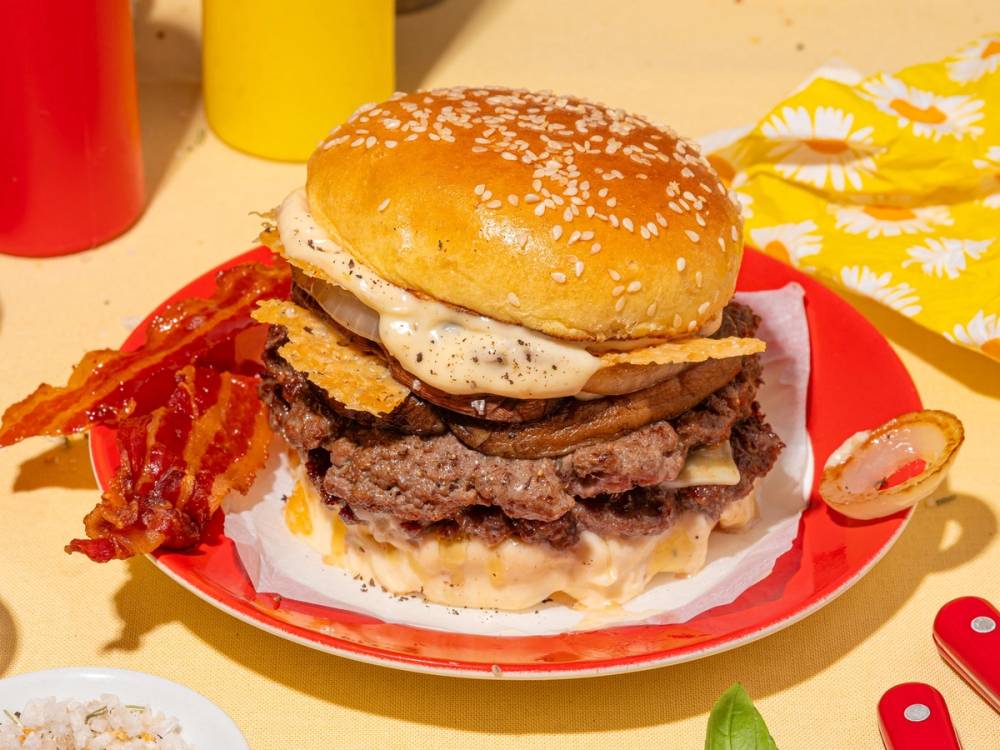 En saftig smashburger med ost, salat og bacon på en rød tallerken, omgitt av ketchup og sennep på bordet.