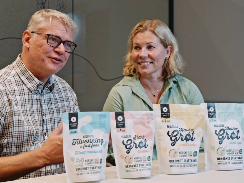 To personer sitter ved et bord med flere poser av den sorte havreproduktet foran seg, og diskuterer noe.