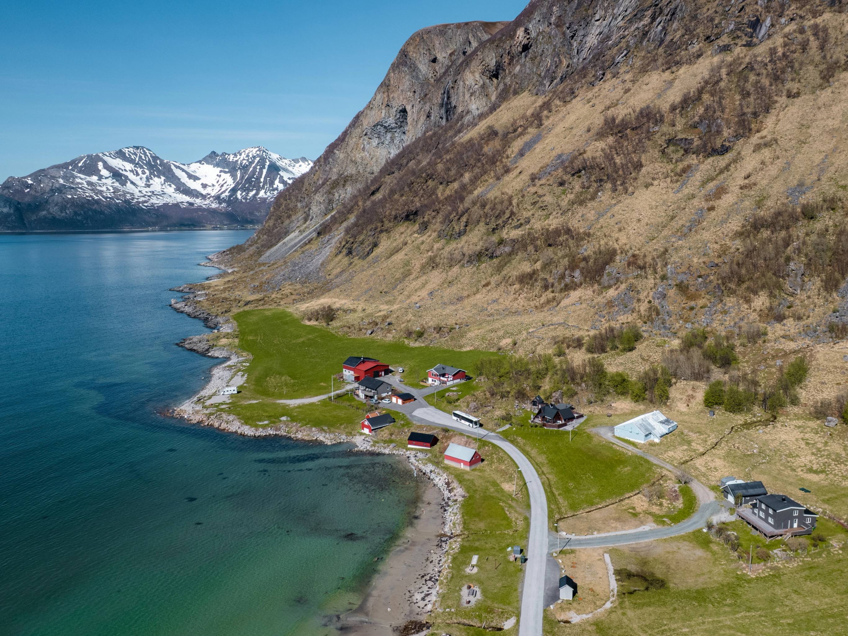En liten landsby ved fjorden med hus og en vei som går langs vannet, omgitt av fjell med snø på toppen.