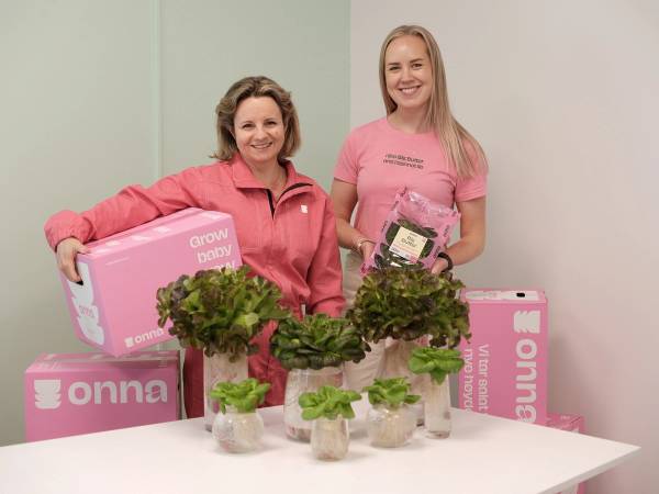 To personer står bak en bord med ulike typer salat, og holder opp pakker med salatprodukter, med en rosa Onna-emballasje i bakgrunnen.