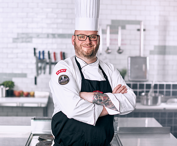 En kok i profesjonell uniform står med armene i kors på et kjøkken med hvite fliser og kjøkkenutstyr i bakgrunnen.