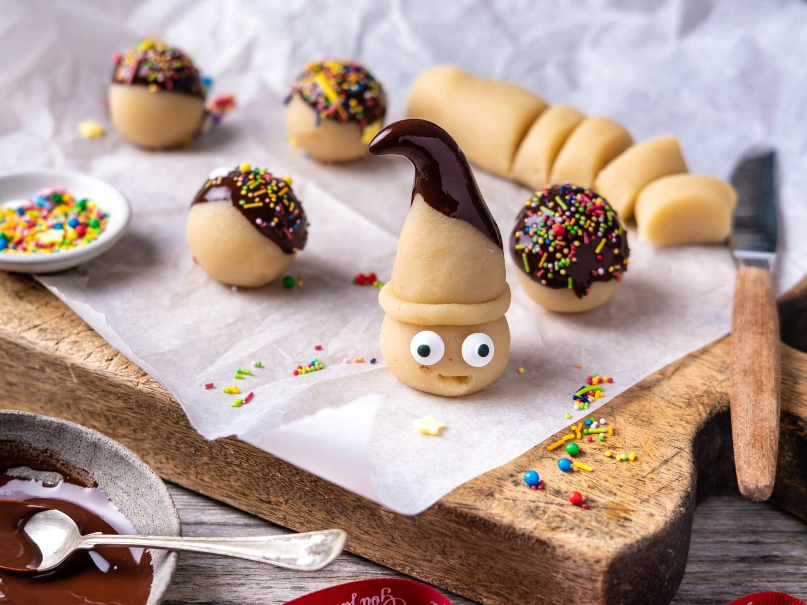 Julemarsipan formet som en larve med fargerike sprinkles på hodet, plassert på et skjærebrett med flere små marsipansnurrer i bakgrunnen.