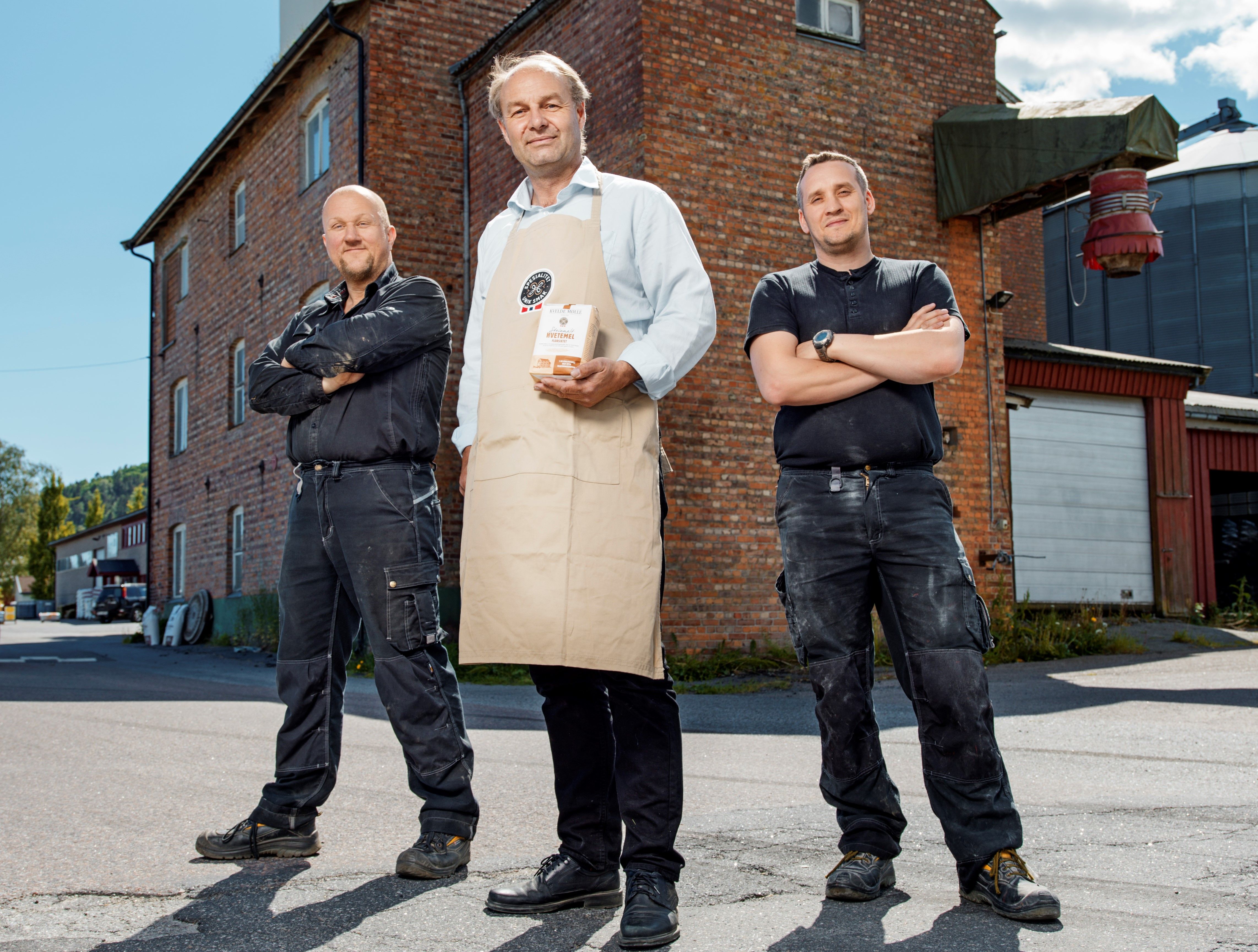 En person i hvit skjorte og beige forkle står mellom to personer i svarte klær med armene i kors foran en rød murbygning, med en bygning og en garasje i bakgrunnen.