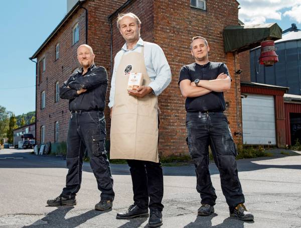 En person i hvit skjorte og beige forkle står mellom to personer i svarte klær med armene i kors foran en rød murbygning, med en bygning og en garasje i bakgrunnen.