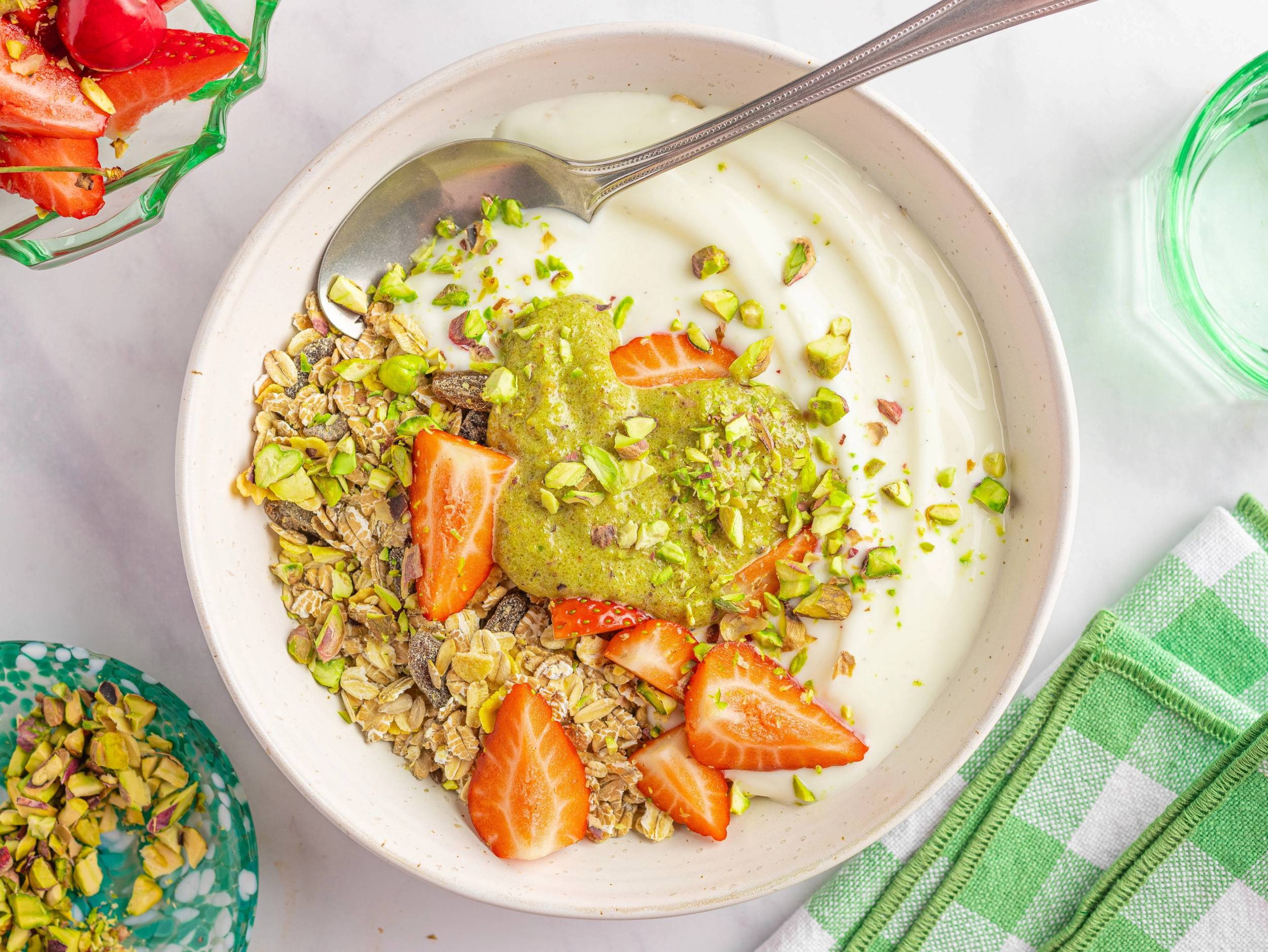 Pistasj krem med jordbær og nøtter i en hvit bolle med en skje, omgitt av en grønn og hvit kjøkkenklut og andre matvarer.
