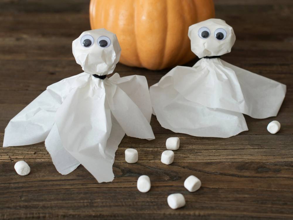 To små spøkelser laget av matpapir med store øyne foran et lite gresskar, omgitt av små hvite marshmallows på en treflate.