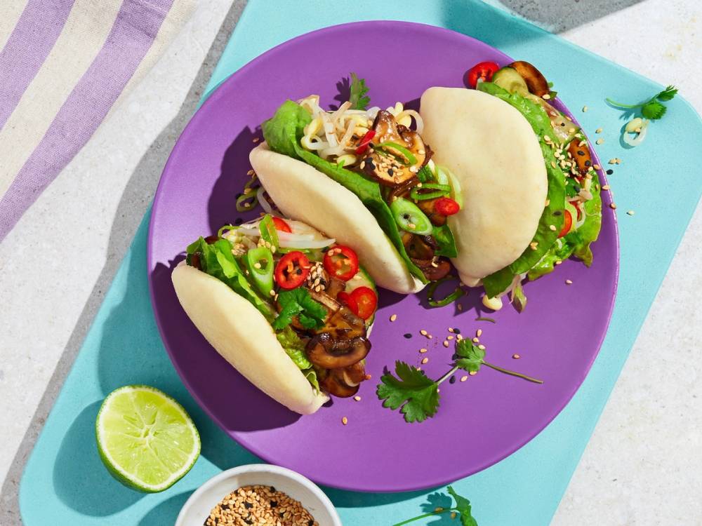 Tre bao buns fylt med kylling, sopp og grønnsaker på en lilla tallerken med lime og krydder ved siden av.