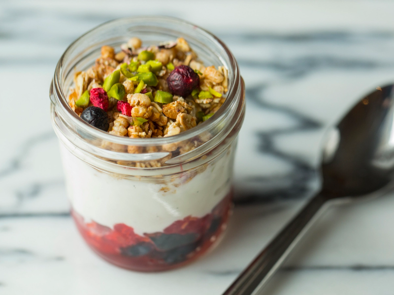 Yoghurt med granola og bær i et glass, plassert på en hvit og grå marmorbord med en skje ved siden av.
