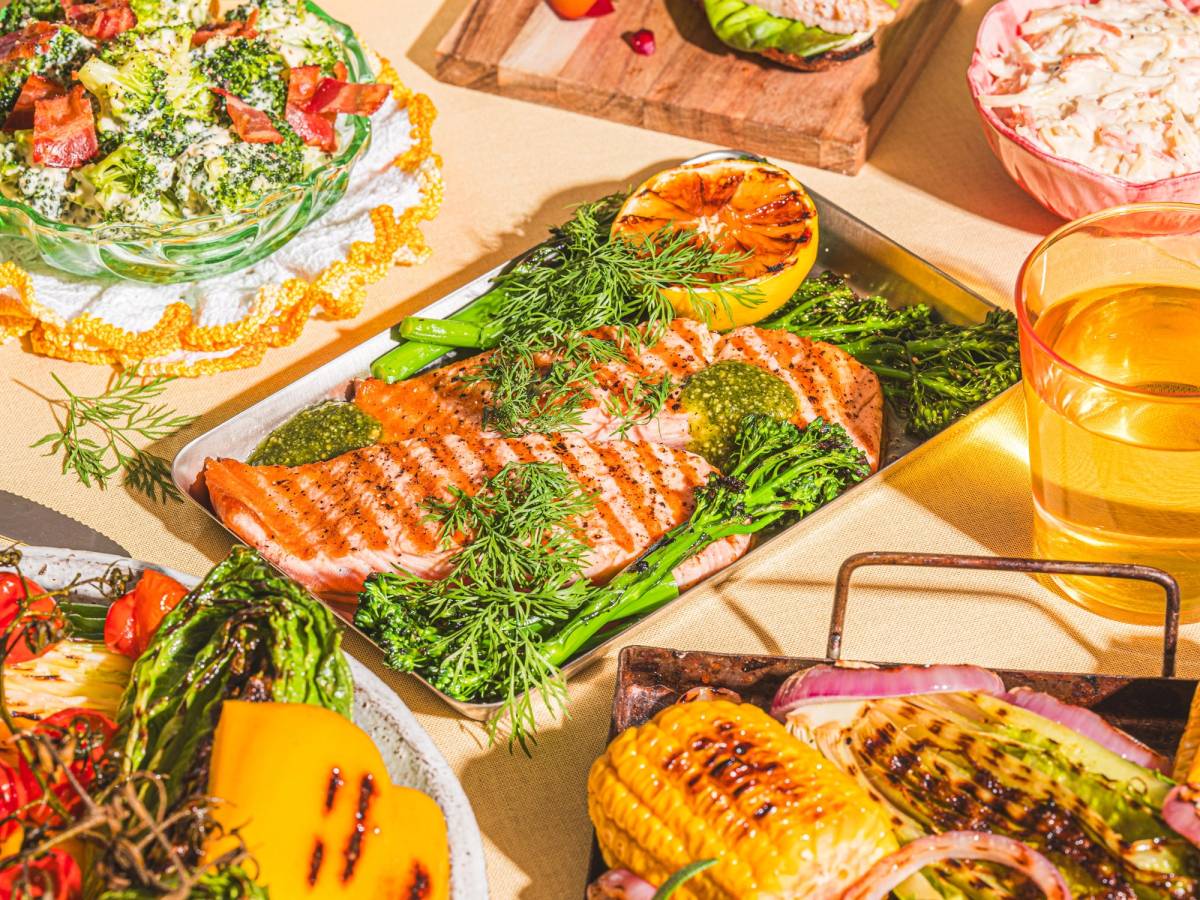 Grillet laks med grønnsaker og urter på et serveringsfat, omgitt av ulike sommerretter og drikkevarer.