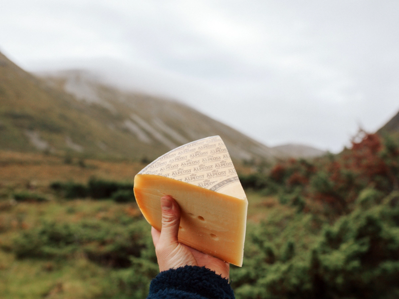 En hånd holder en skåret alpeost mot et fjellandskap med fjell og åpen himmel i bakgrunnen.