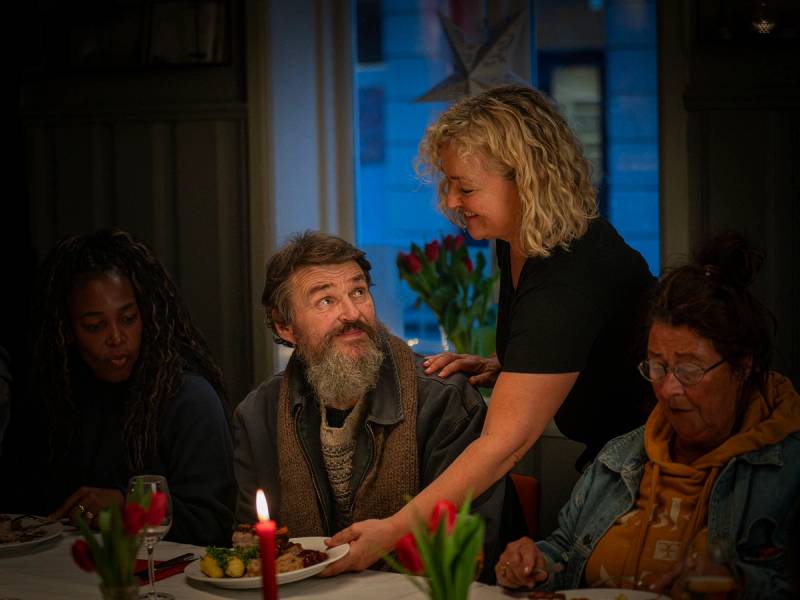En kvinne serverer mat til en gruppe mennesker rundt et bord med lys og blomster, sannsynligvis i en sosial eller veldedighetssetting.