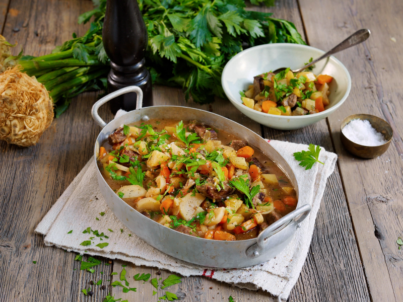 Lapskaus i en stor gryte med grønnsaker og kjøtt, omgitt av ferske urter og krydder på et kjøkkenbord.