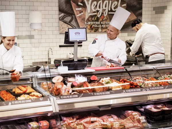 Kjøkkenpersonell jobber bak disken på en matbutikk, med en meny-liste for Fornebu synlig i bakgrunnen.