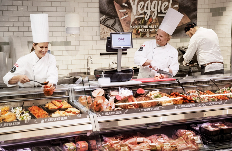Kjøkkenpersonell jobber bak disken på en matbutikk, med en meny-liste for Fornebu synlig i bakgrunnen.