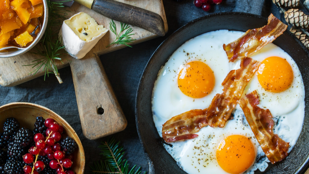 Tre stekte egg med bacon på en svart panne, omgitt av frukt og urter på et frokostbord.