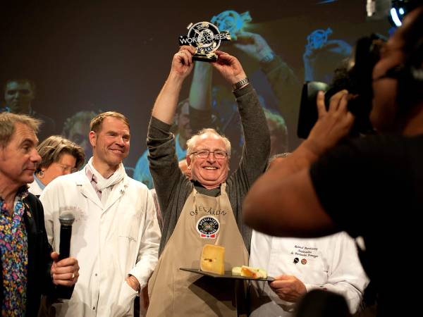 En person holder opp en pokal på en premieutdeling med flere andre personer rundt seg, noen med mikrofon og kamera, i en innendørs setting.