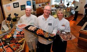 Tre ansatte i hvite uniformer står bak en serveringsdisk med ulike retter i en restaurant eller kafé, med kunder i bakgrunnen.