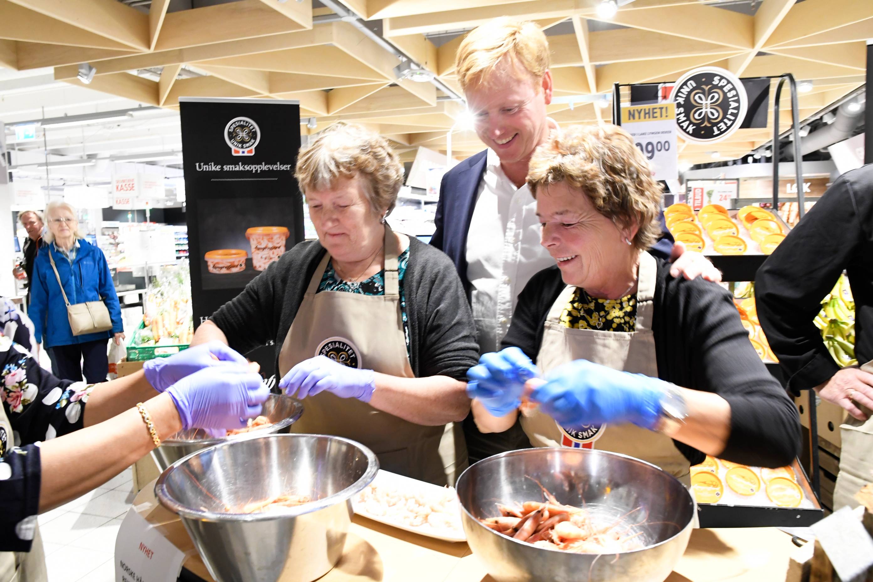 Personer som deltar i rekepilling under en demonstrasjon i en butikk, med en person som har på seg en hvit kokkejakke og andre med blå hansker, mens en annen person holder en bolle med rekeskall.