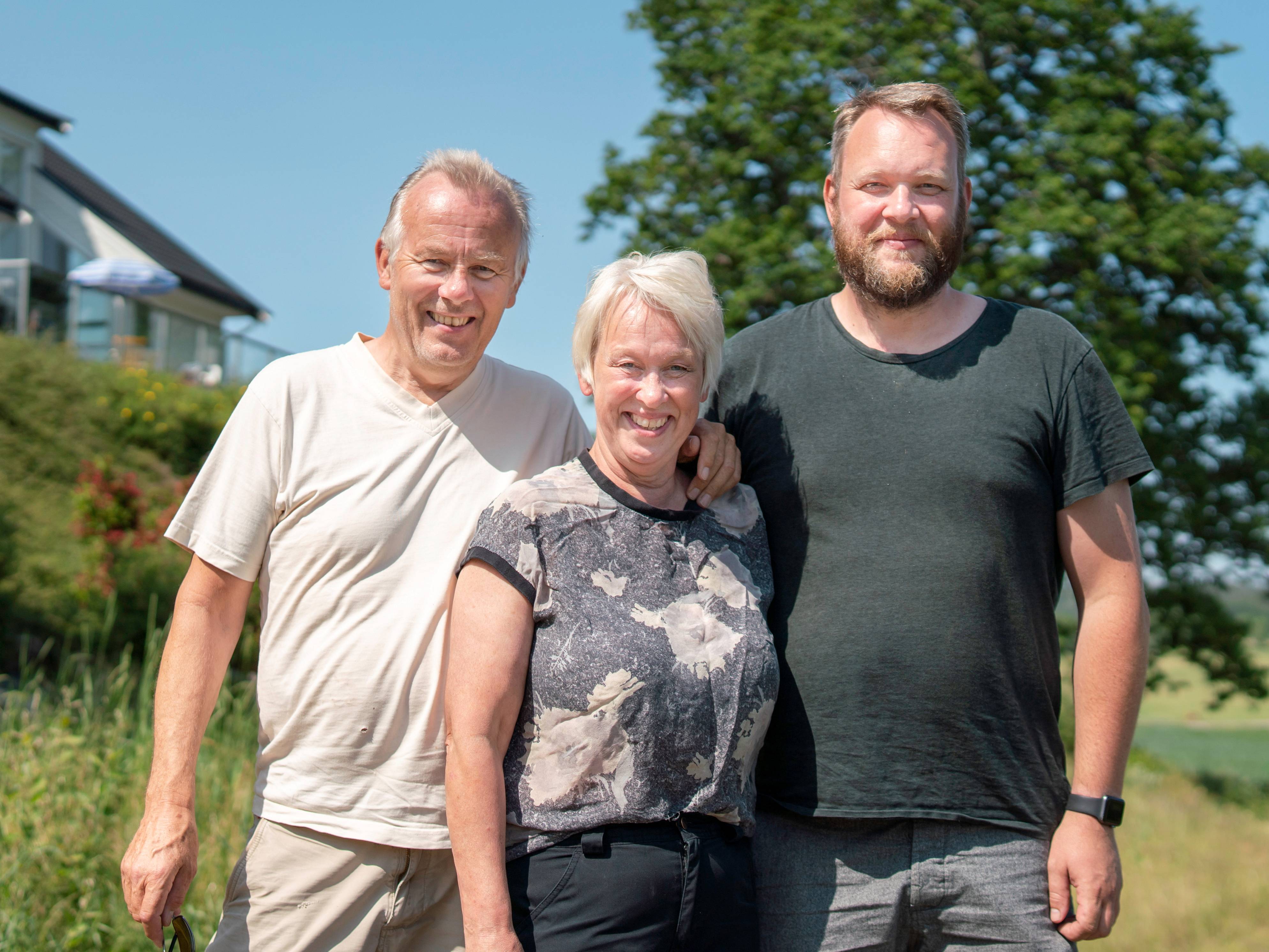 Tre personer står ute i solskinn med grønne trær og hus i bakgrunnen, alle med ansiktene blurred.