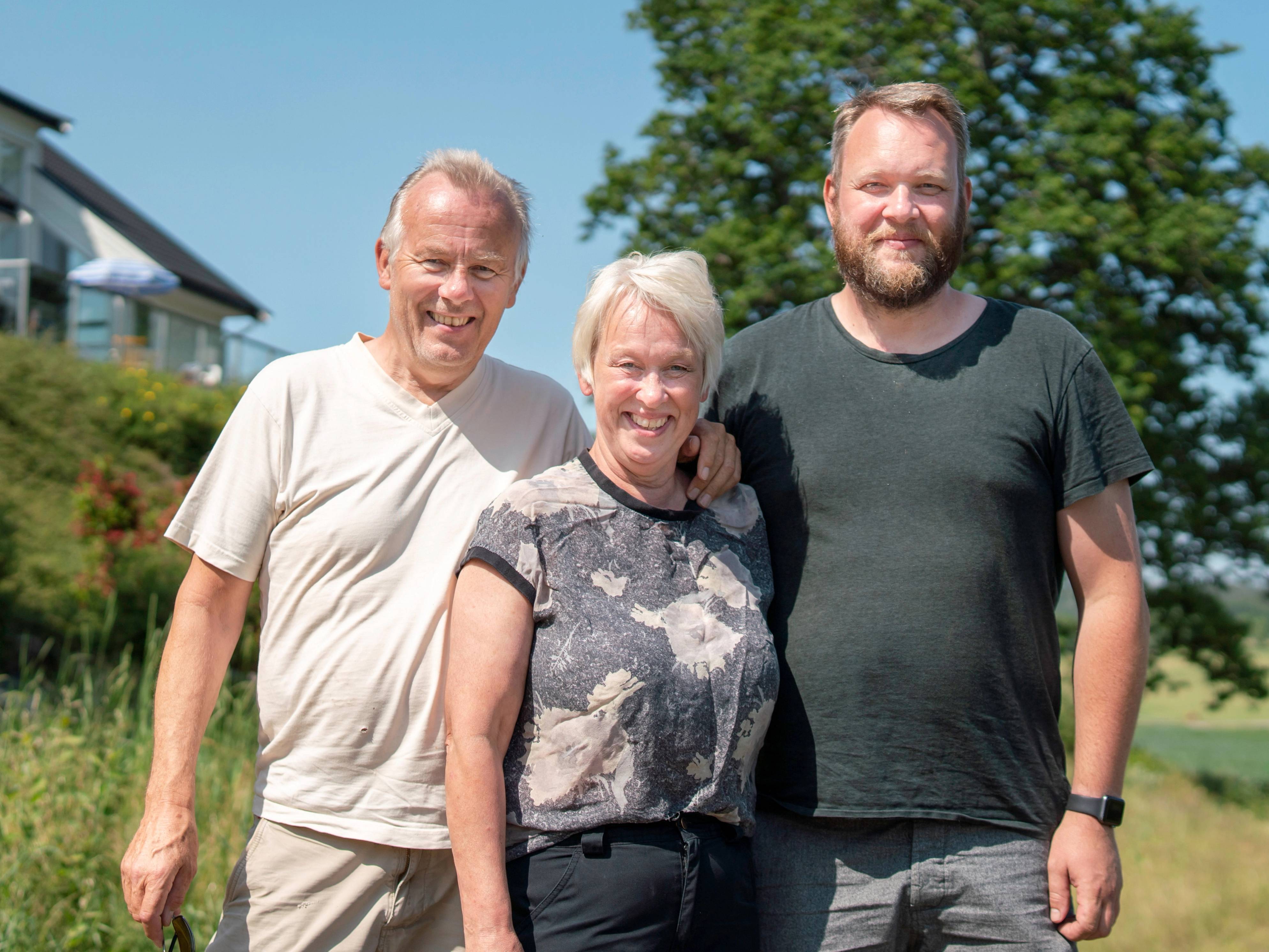 Tre personer står ute i solskinn med grønne trær og hus i bakgrunnen, alle med ansiktene blurred.