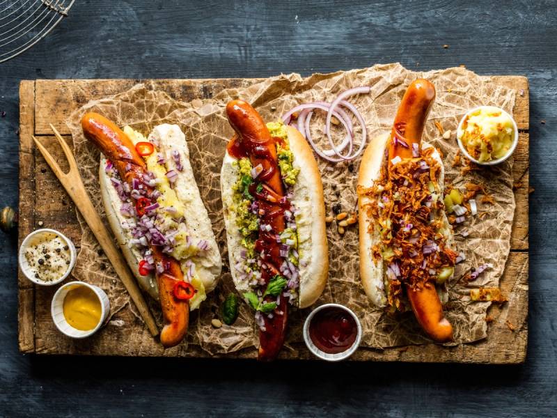 Tre pølser i brød med ulike toppings på et trestykke papir, omgitt av små skåler med tilbehør.