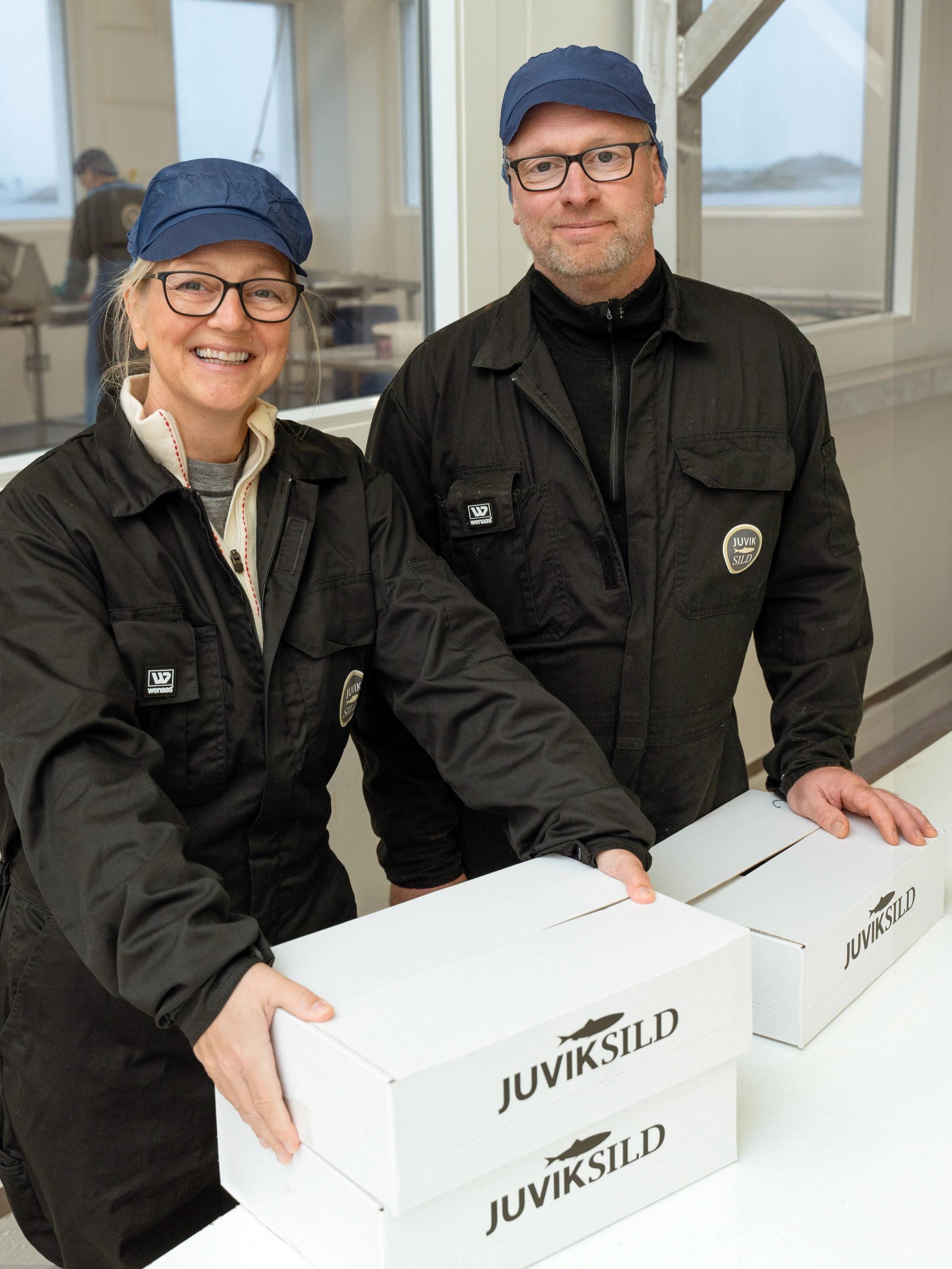 To personer står ved et bord med to Juviksild-kasser, og de holder hver sin kasse, mens de ser mot kameraet, i et rom med store vinduer.