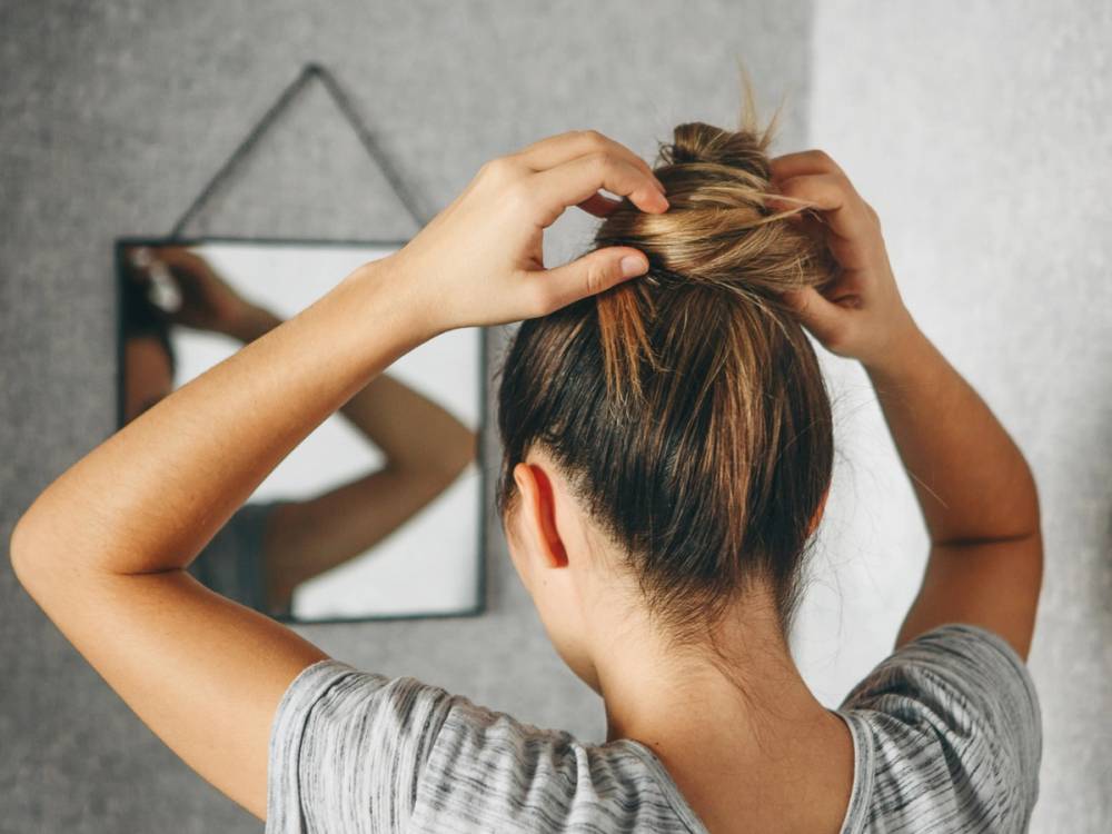 En person med oppsatt hår som bruker hendene til å justere frisuren, sett fra bak, med et speil i bakgrunnen.