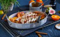 Kalkunbryst skåret opp og servert med sitronbåter i en stor metallform, omgitt av urter og krydder på et mørkt bord.