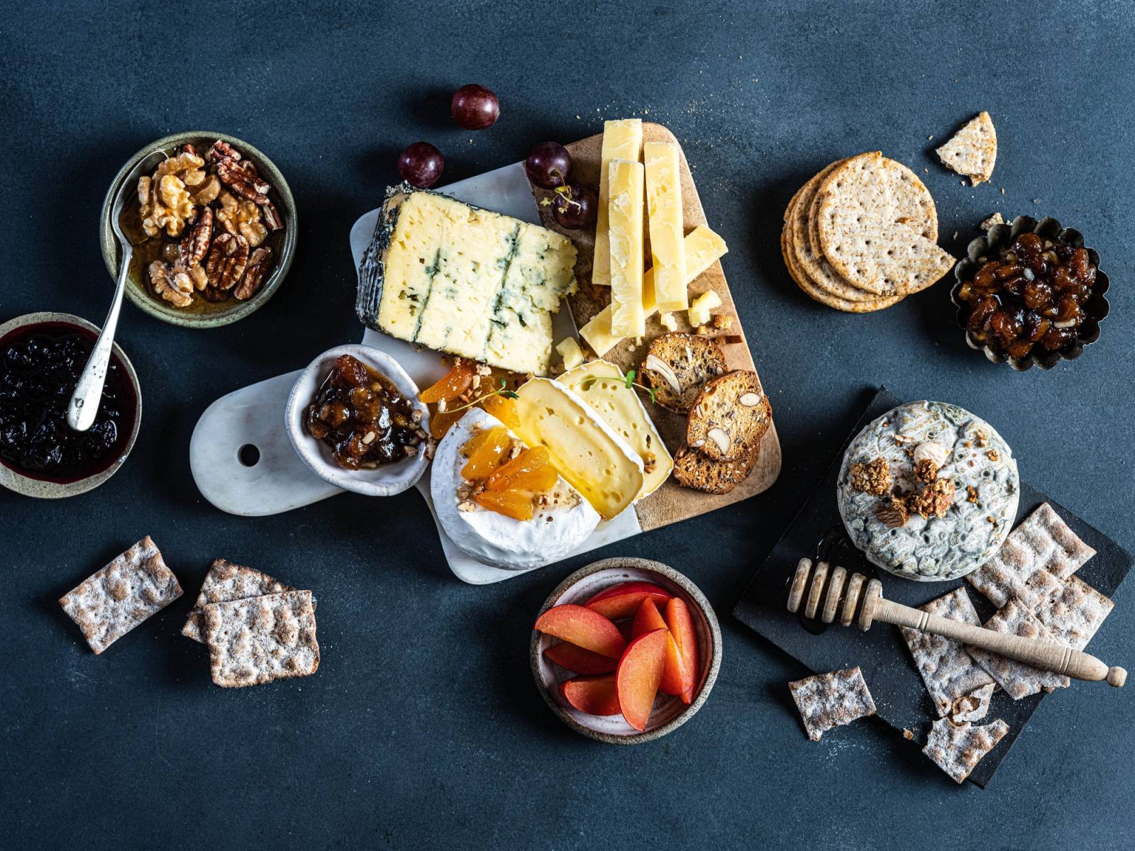 Et utvalg av oster, kjeks, nøtter og tørket frukt på et mørkt bord.