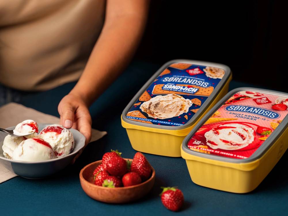 To isbokser med Sørlandschips på et bord, en skål med jordbær og en person som holder en skål med is med jordbær og krem, delvis synlig i bakgrunnen.