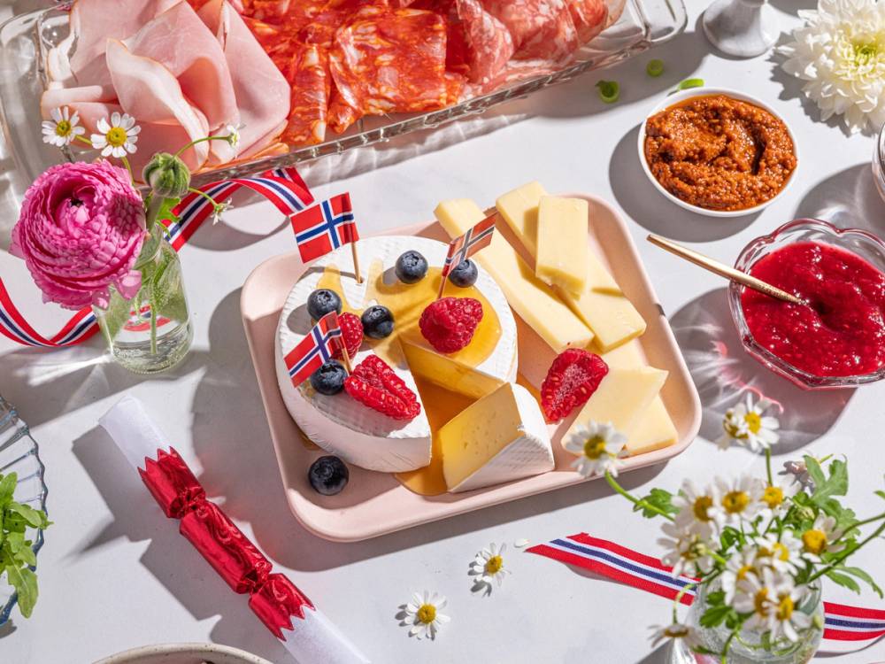 Utsikt av en 17. mai frokost med ost, bær, kjeks og tilbehør på et bord pyntet med norske flagg og blomster.