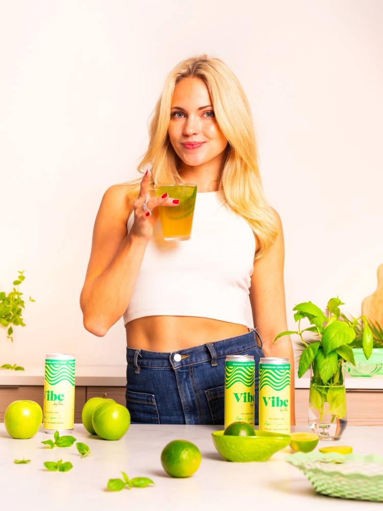 En kvinne med lang blond hår holder et glass med grønn drikke, omgitt av grønne epler og Vibe By Voe-19 produkter på kjøkkenbenken.