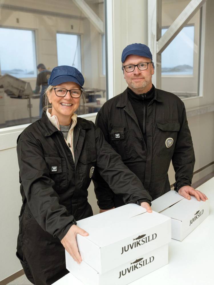 To personer i arbeidsklær står ved et bord med to hvite bokser merket Juviksild, i et rom med store vinduer og andre personer i bakgrunnen.