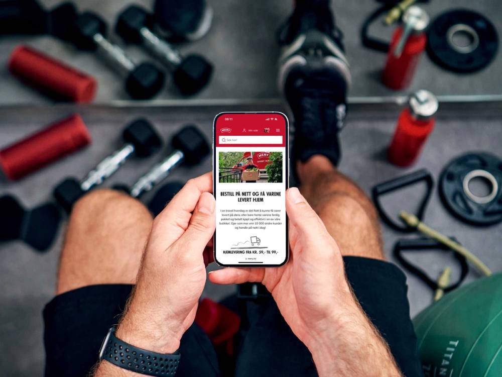 En person som bruker en mobiltelefon med en meny for levering hjem, omgitt av treningsutstyr på gulvet.