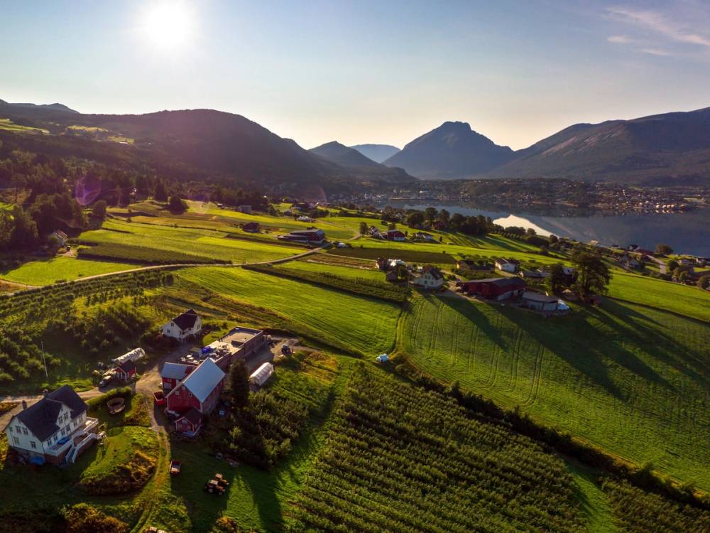 Landskap med grønne jordbruksområder, små hus og fjell i bakgrunnen under en klar himmel.