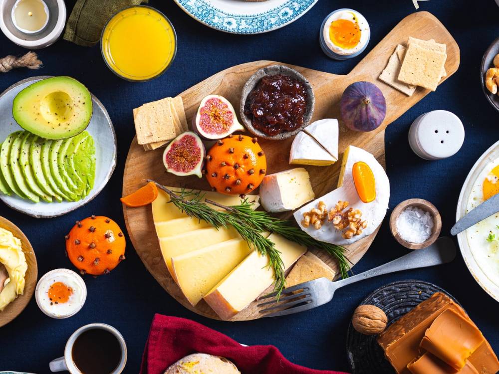 Et julefrokostbord med vegetariske oster, frukt, kjeks og pålegg, sett på et mørkt bord med ulike småretter og drikkevarer.